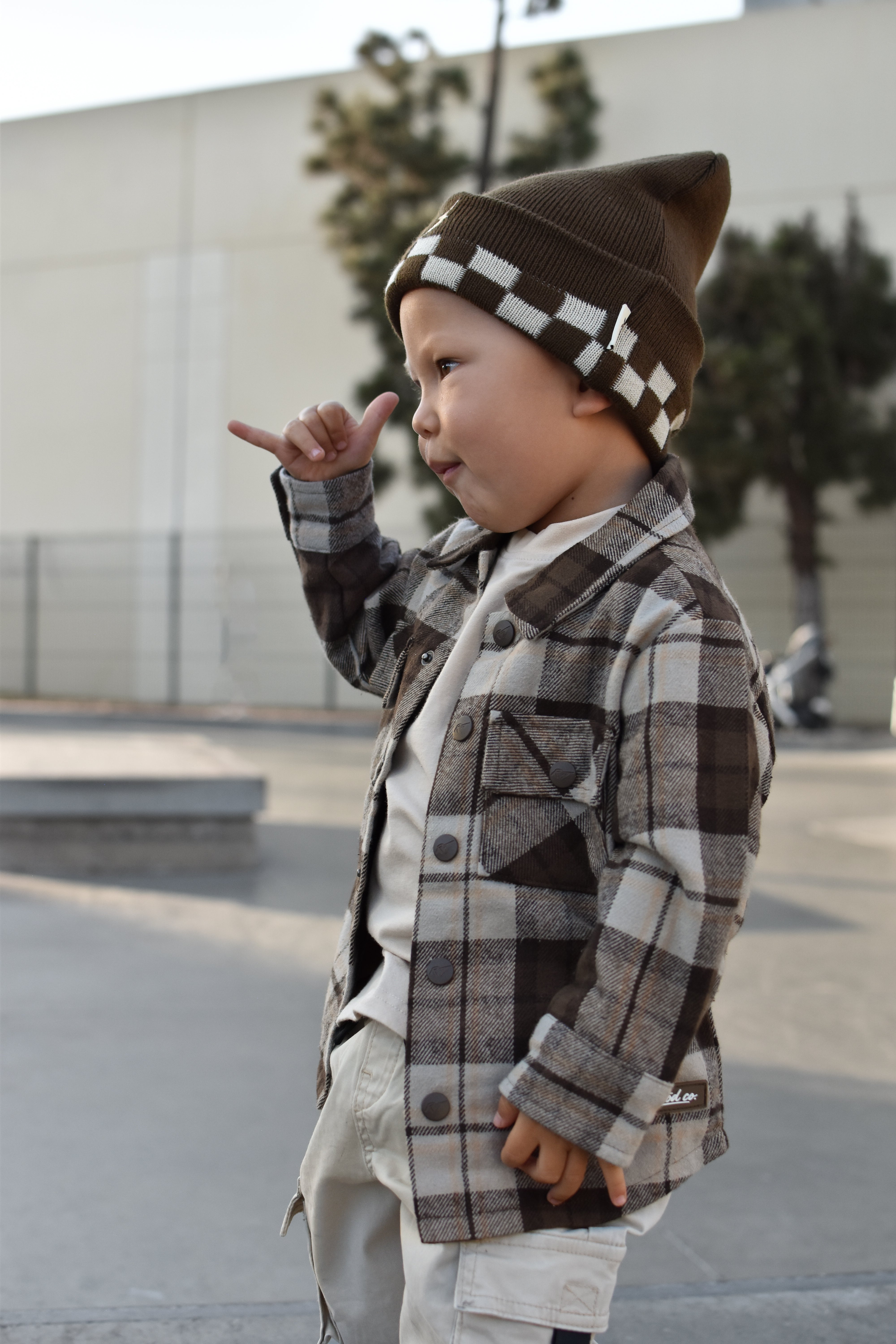 A toddler standing outdoors wearing a brown and beige plaid button-up jacket over a white shirt, paired with a brown knit beanie featuring a white checkerboard pattern.