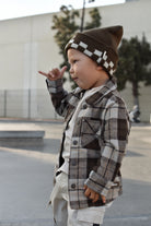 A toddler standing outdoors wearing a brown and beige plaid button-up jacket over a white shirt, paired with a brown knit beanie featuring a white checkerboard pattern.