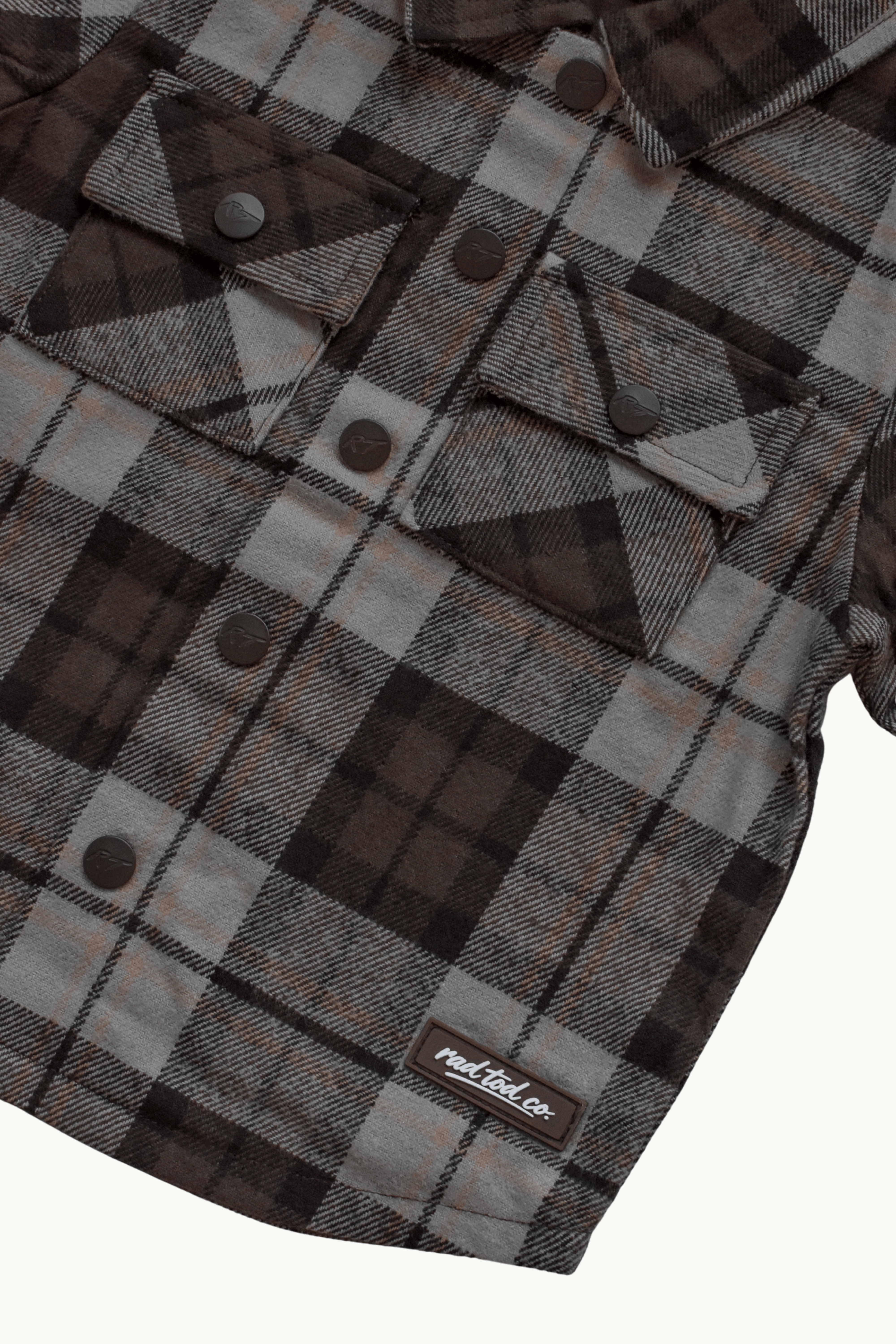 A close-up of a plaid flannel shirt jacket in brown, gray, and black, featuring snap-button chest pockets and a visible 'RAD TOD co.' label near the bottom.