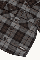A close-up of a plaid flannel shirt jacket in brown, gray, and black, featuring snap-button chest pockets and a visible 'RAD TOD co.' label near the bottom.