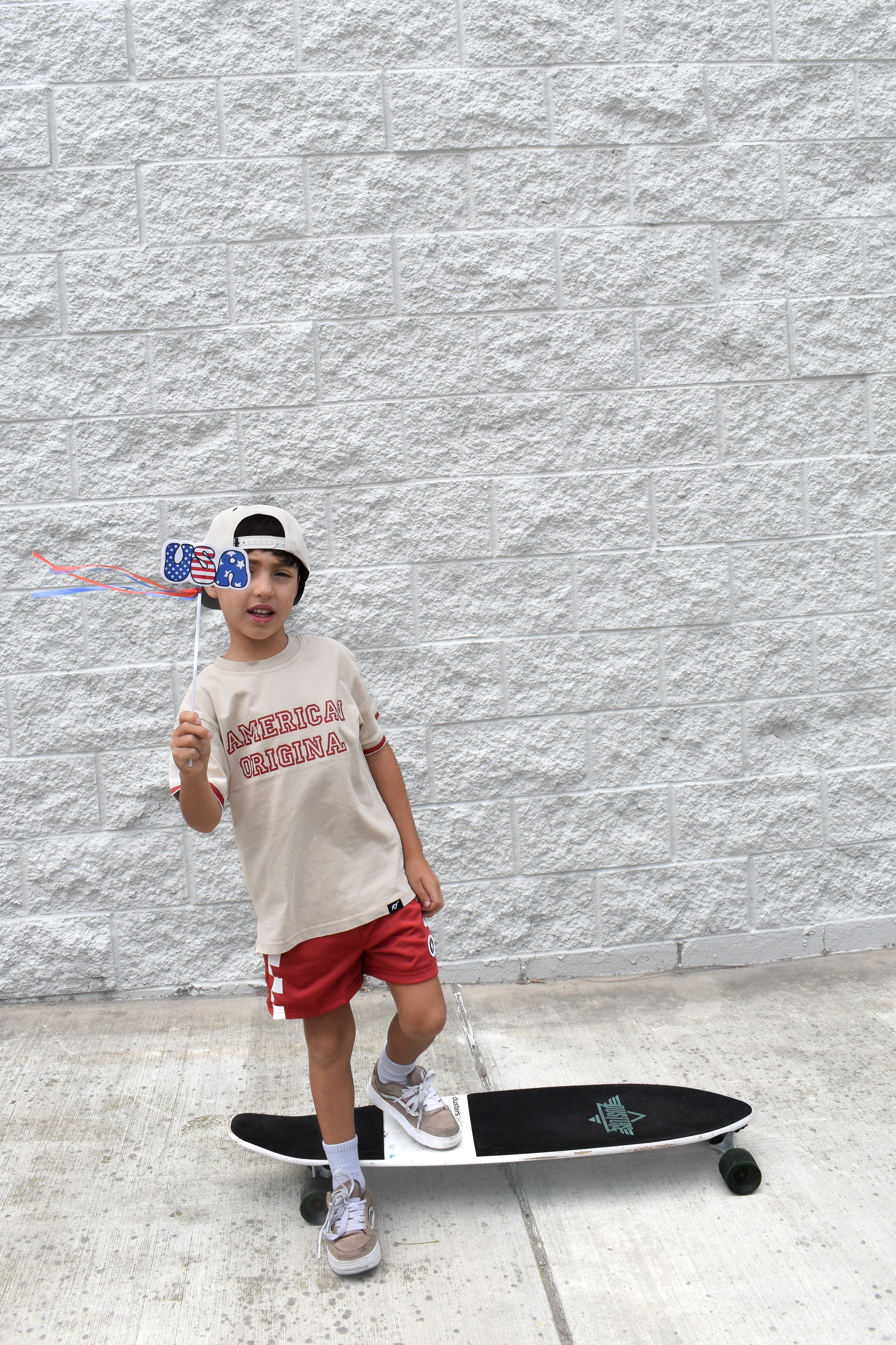 Child with a skateboard and American flag hat standing against a gray wall. Shirt by Rad Toddler