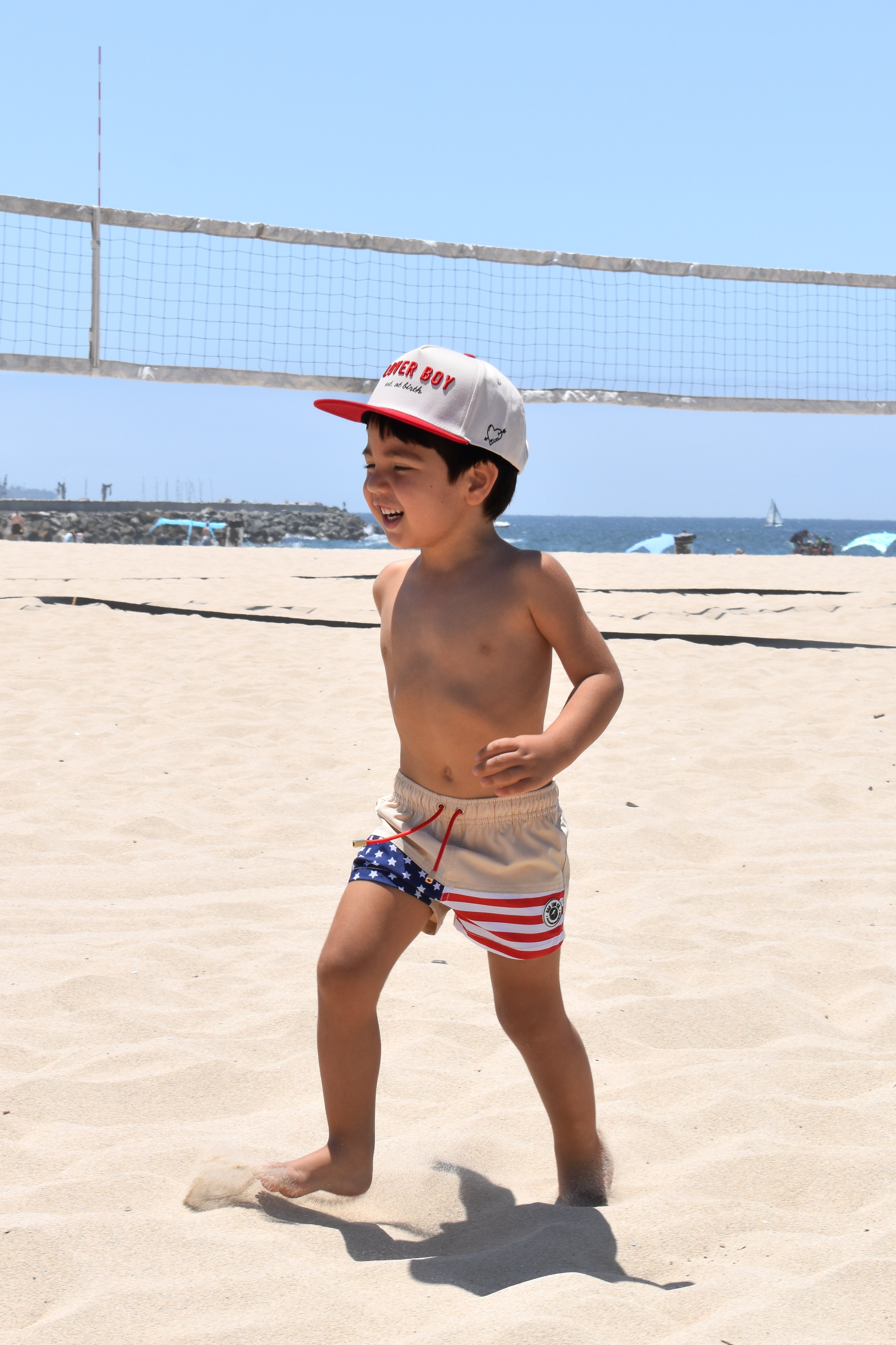 Child in swim trunks and cap on a sandy beach with ocean view