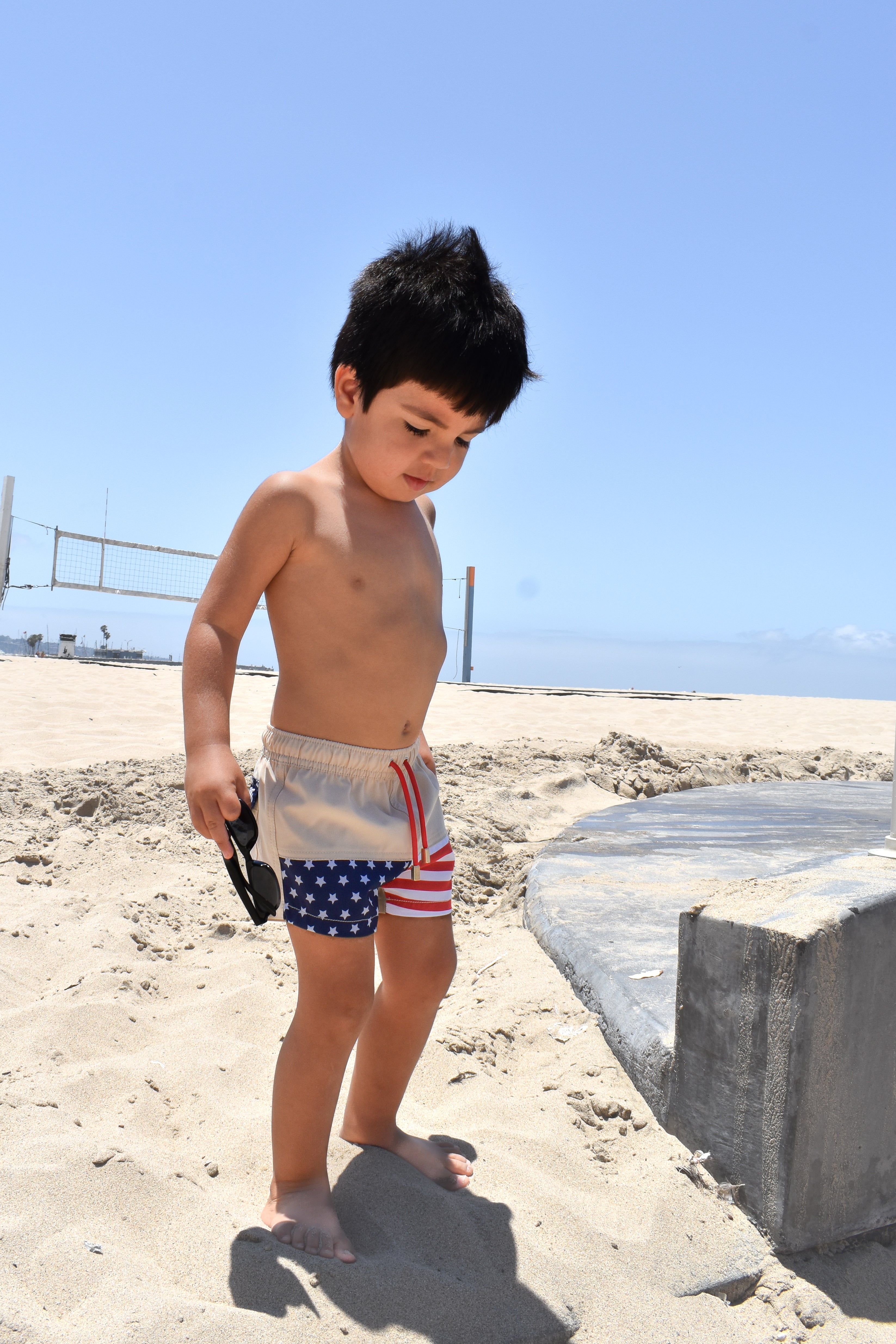 Child in American flag swim shorts standing on a sandy beach with a clear blue sky.