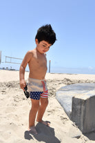 Child in American flag swim shorts standing on a sandy beach with a clear blue sky.