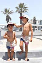 Two children in matching Americana swimsuits and Loverboy hats by RAD TODDLER standing on a concrete surface with palm trees and buildings in the background.