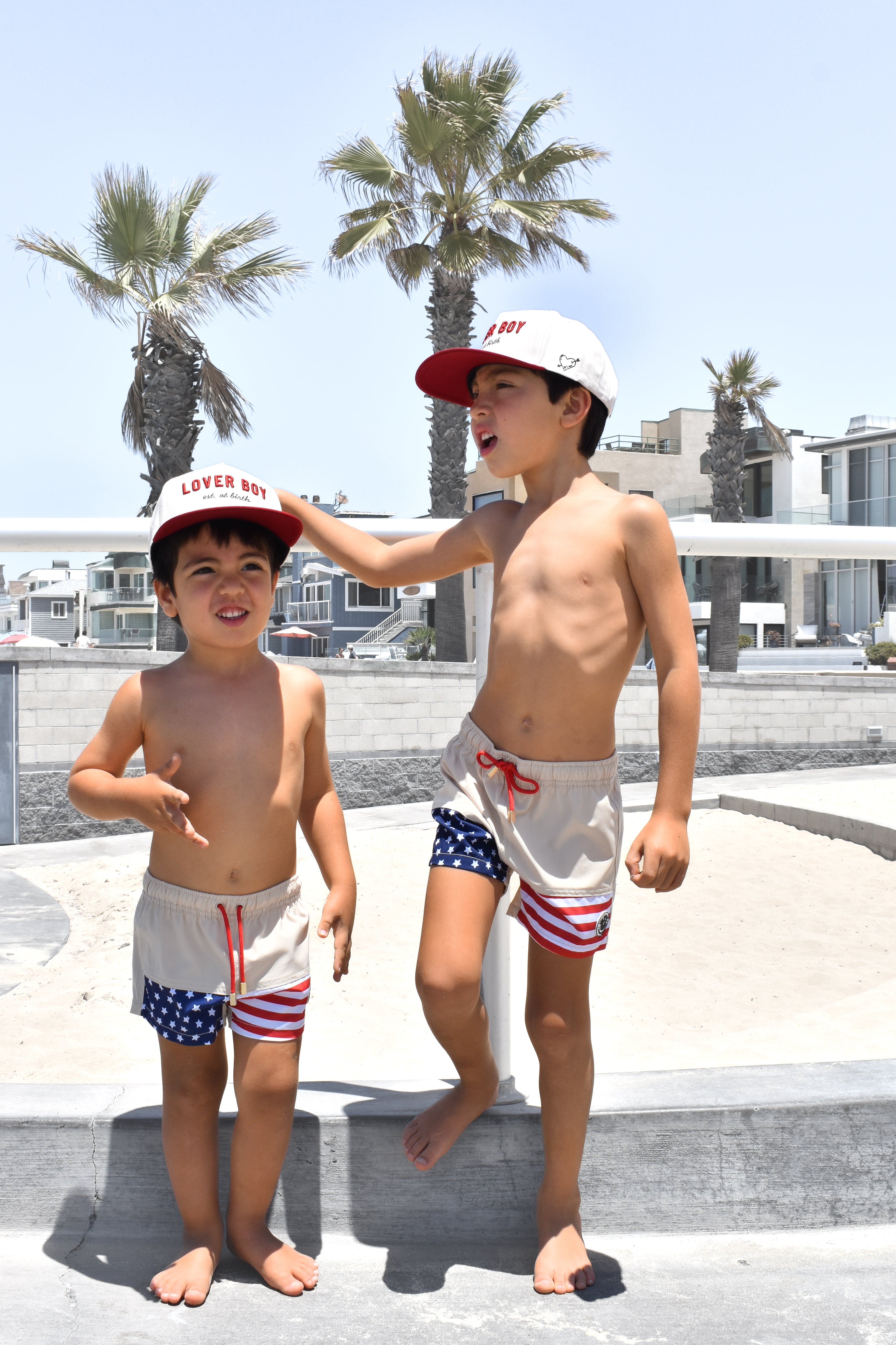 Two children in matching Americana swimsuits and Loverboy hats by RAD TODDLER standing on a concrete surface with palm trees and buildings in the background.