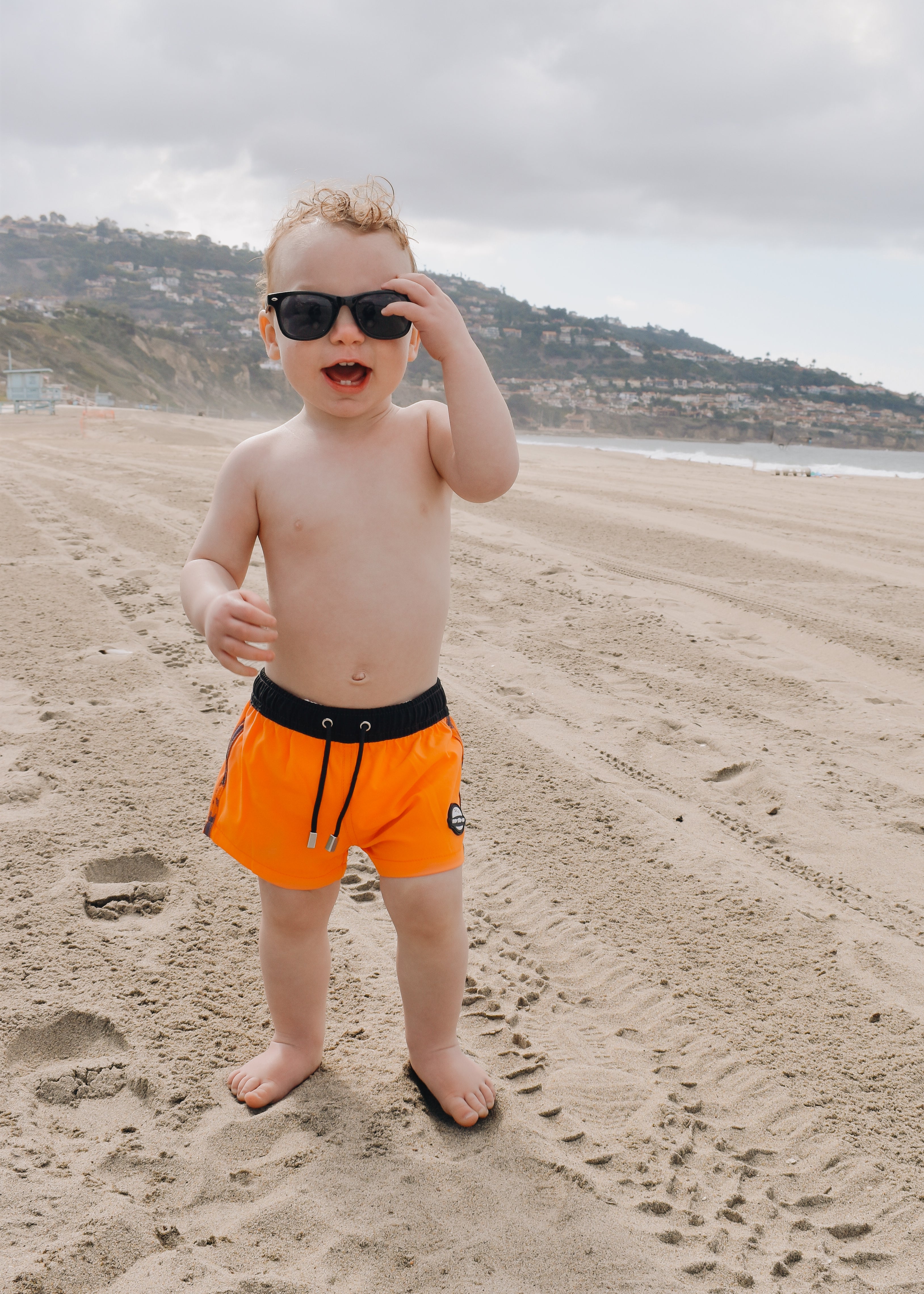 cute beach toddler wearing Bright swim shorts with an elastic waistband and drawstring, made from quick-drying fabric. Ideal for swimming, beach trips, and outdoor activities. Features vibrant patterns and a comfortable fit for active kids.
