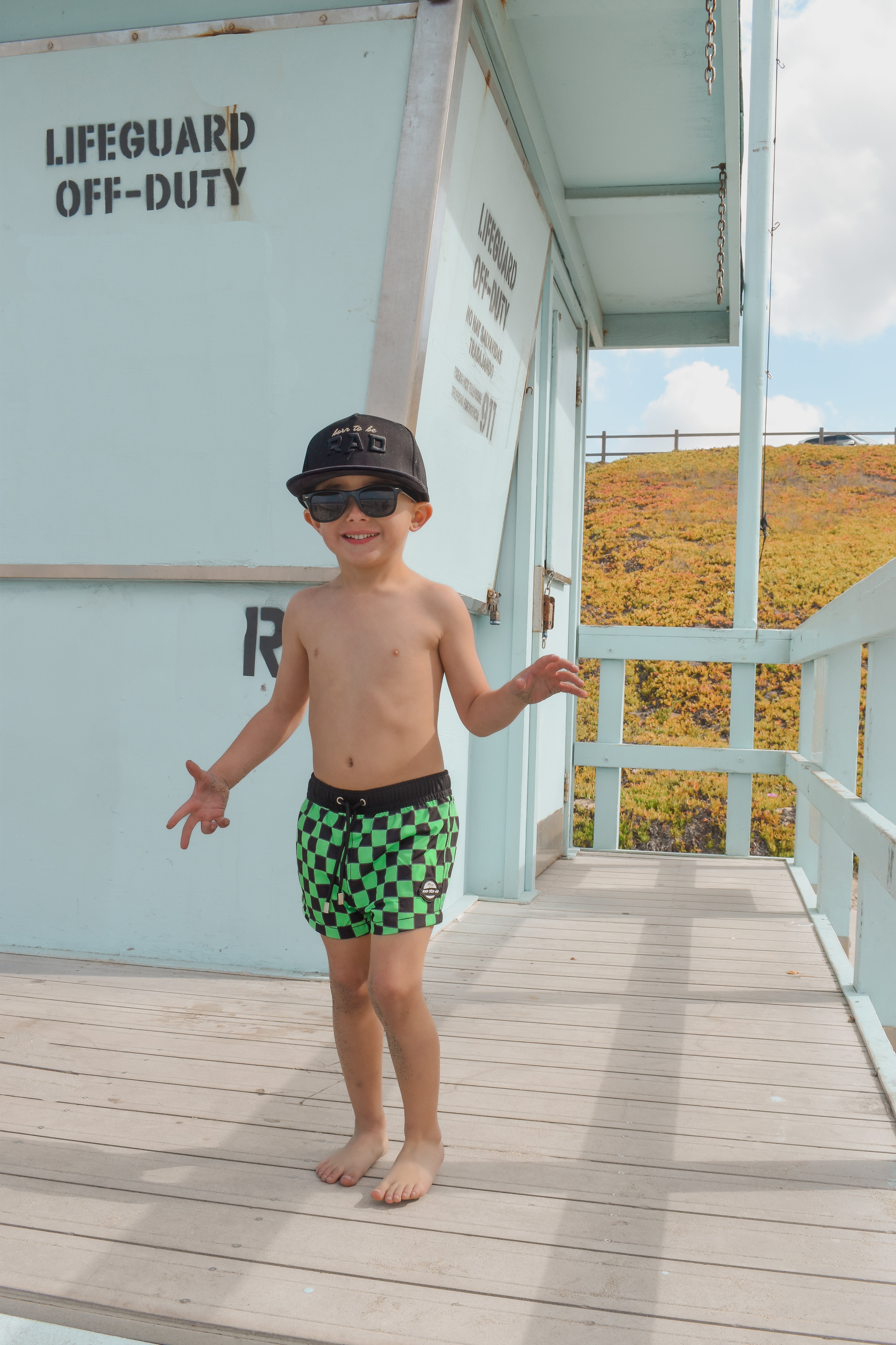 boy on lifeguard tower wearing Bright swim shorts with an elastic waistband and drawstring, made from quick-drying fabric. Ideal for swimming, beach trips, and outdoor activities. Features vibrant patterns and a comfortable fit for active kids.