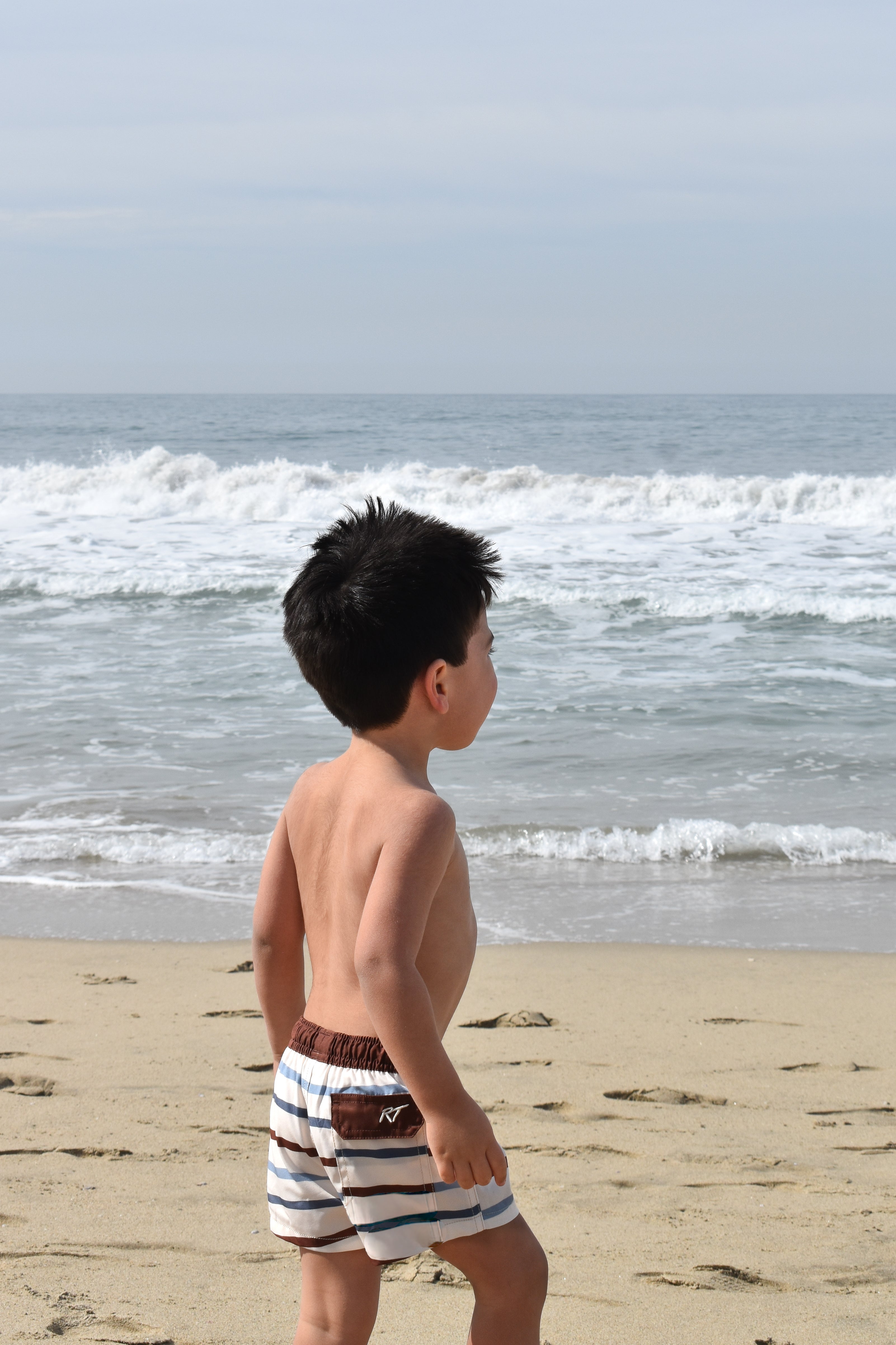 Young boy standing on a beach looking at the ocean wearing Driftwood Stripes by rad toddler