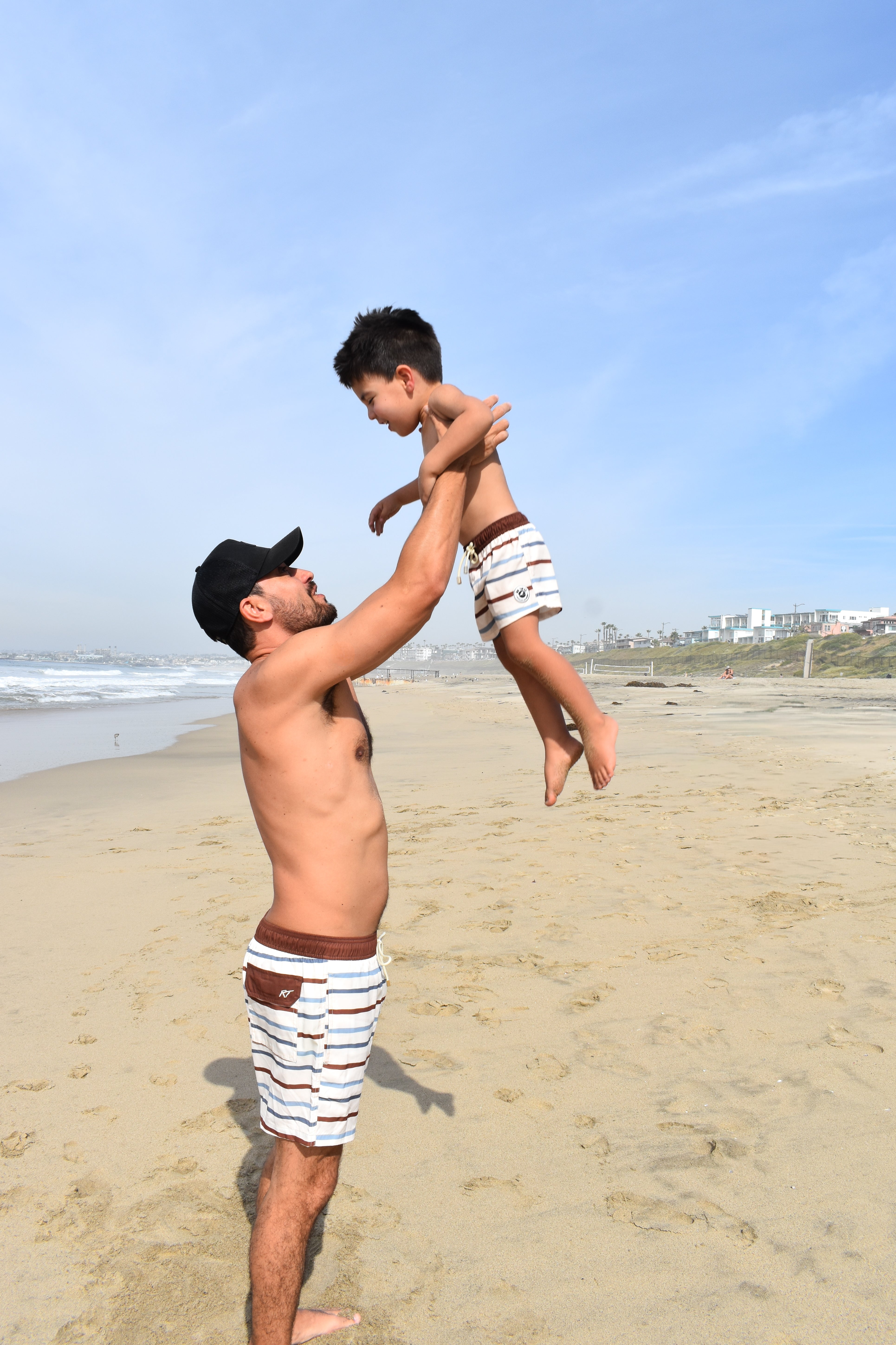 Man lifting a child in the air on a beach with clear blue sky wearing matching Driftwood Stripes swimmies by rad toddler