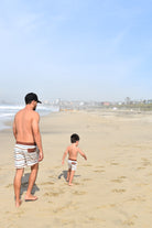 Man and child walking on a sandy beach with a clear sky wearing Driftwood Stripes matching swimmies by rad toddler
