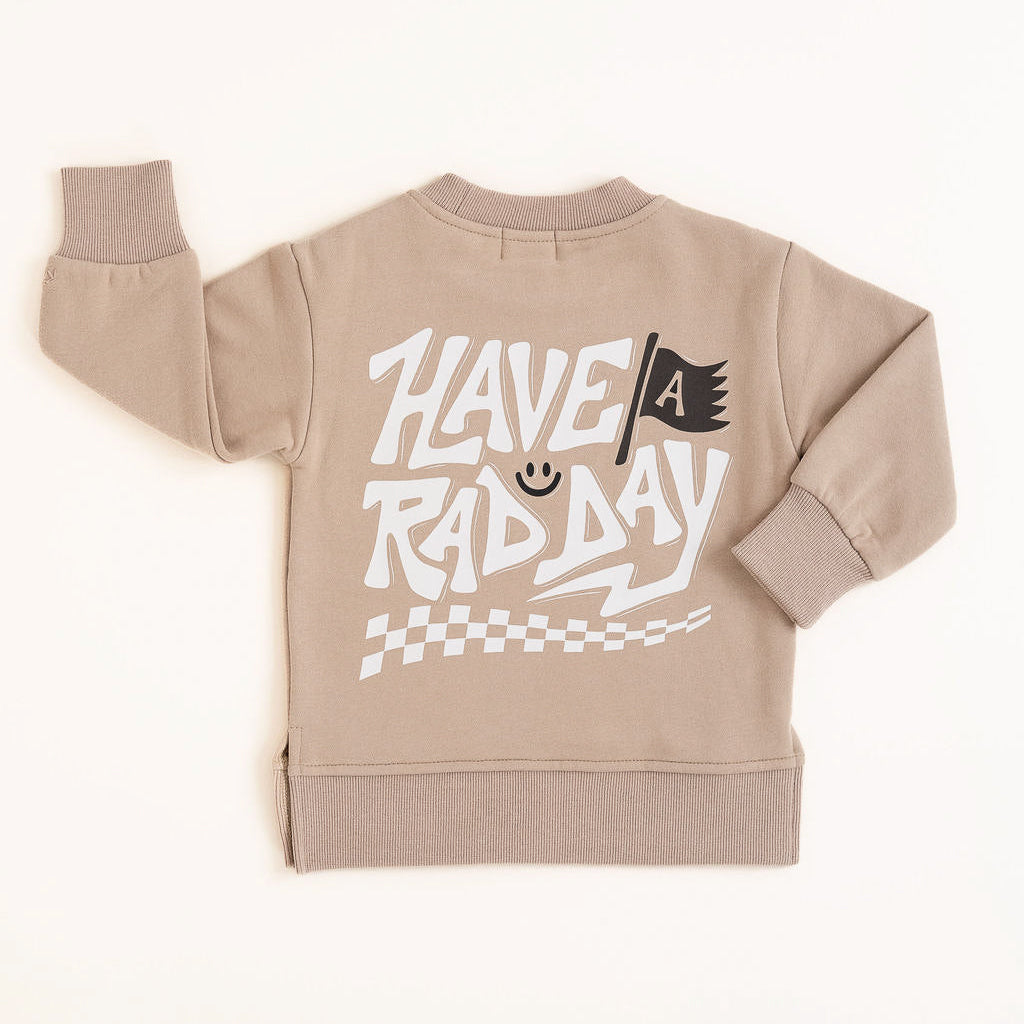 Beige crewneck sweatshirt with white ‘Have a Rad Day’ graphic, flag, smiley face, and checker details laid flat”