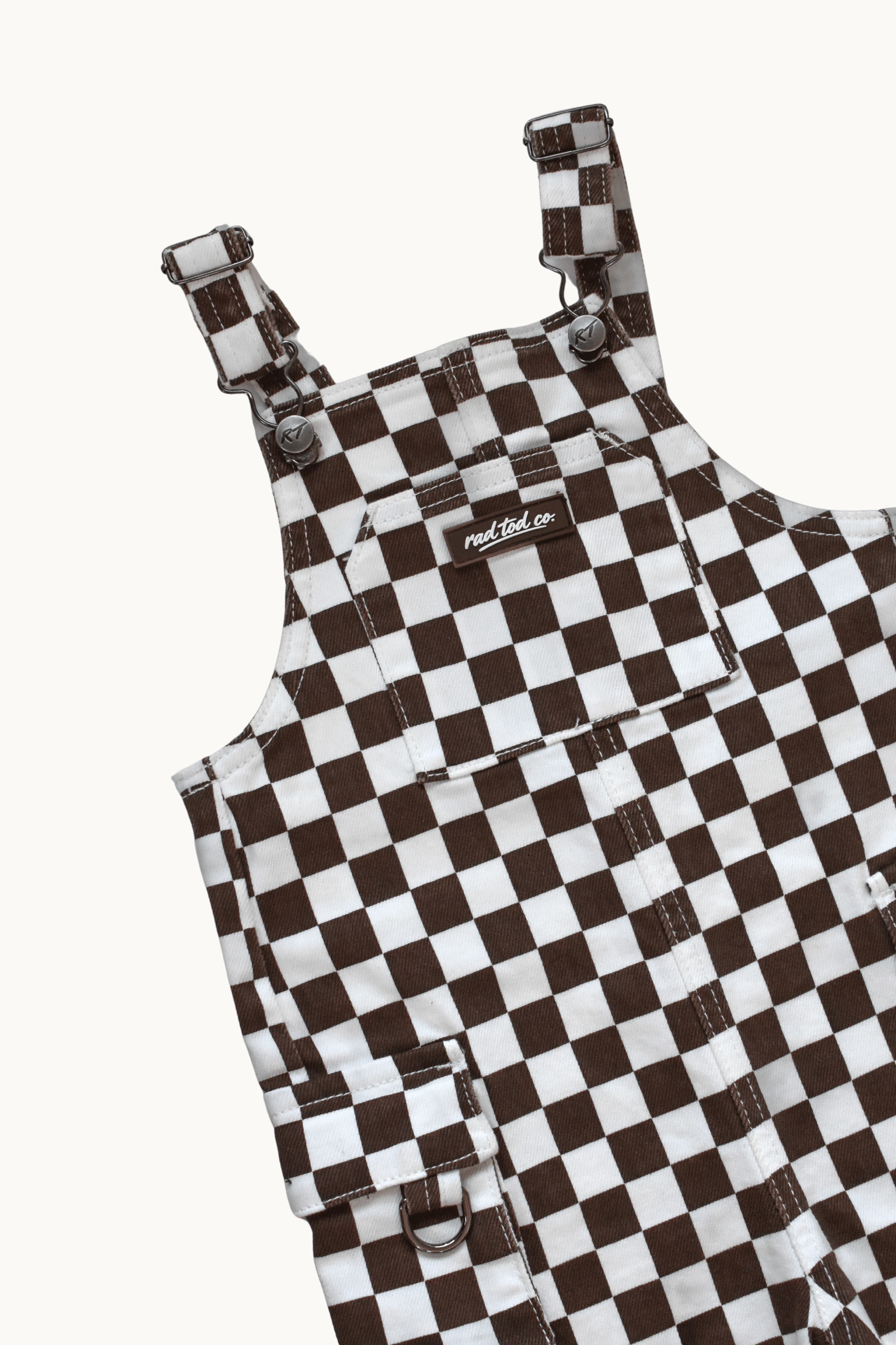 A close-up of a brown and white checkerboard patterned overall garment, showing the shoulder straps, front pockets, and a small visible Rad Tod brand label.