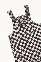 A close-up of a brown and white checkerboard patterned overall garment, showing the shoulder straps, front pockets, and a small visible Rad Tod brand label.