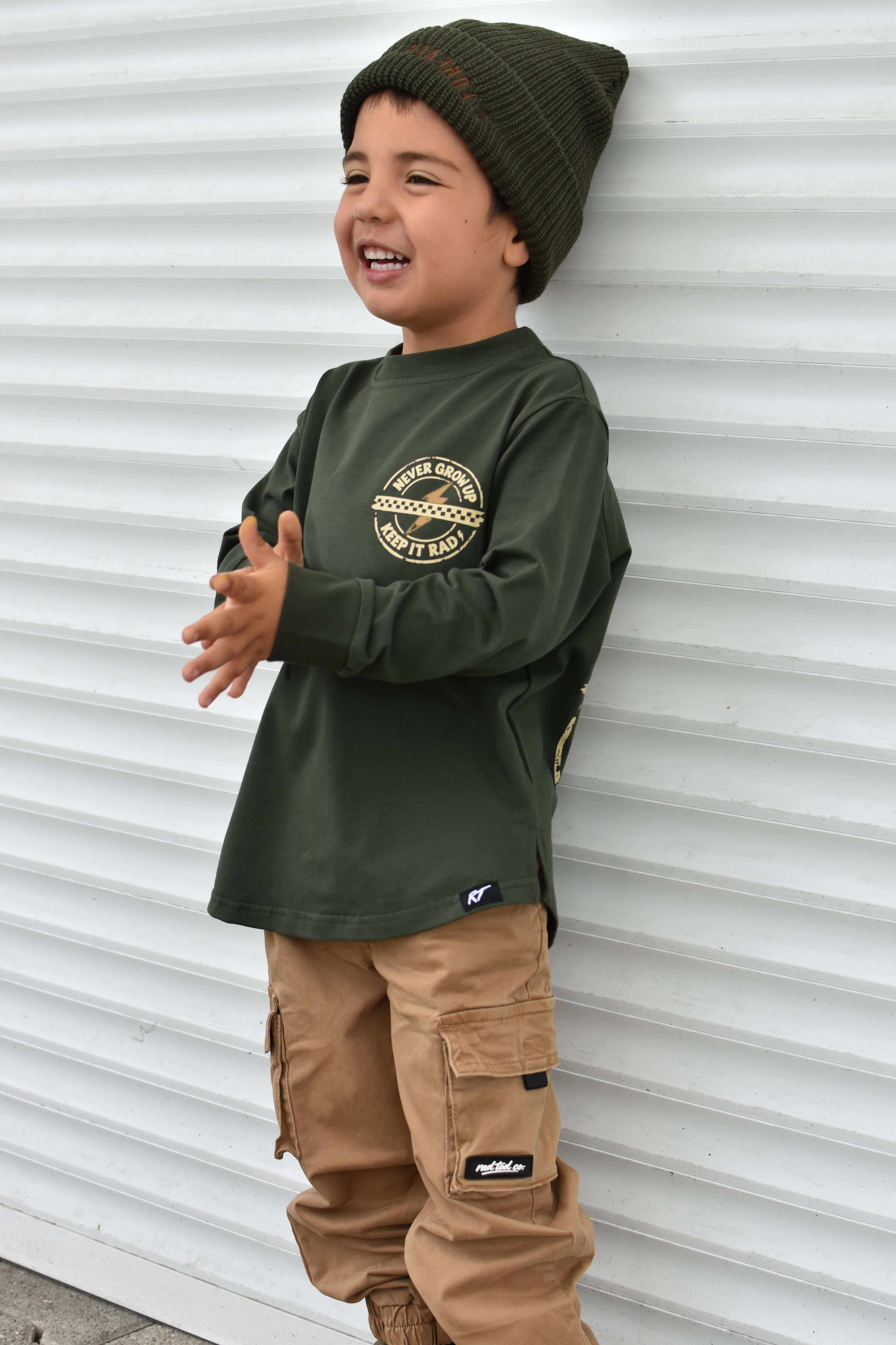 A young boy wearing an olive green long-sleeve graphic shirt, tan cargo pants, and a dark green knit beanie, standing against a white corrugated metal wall.