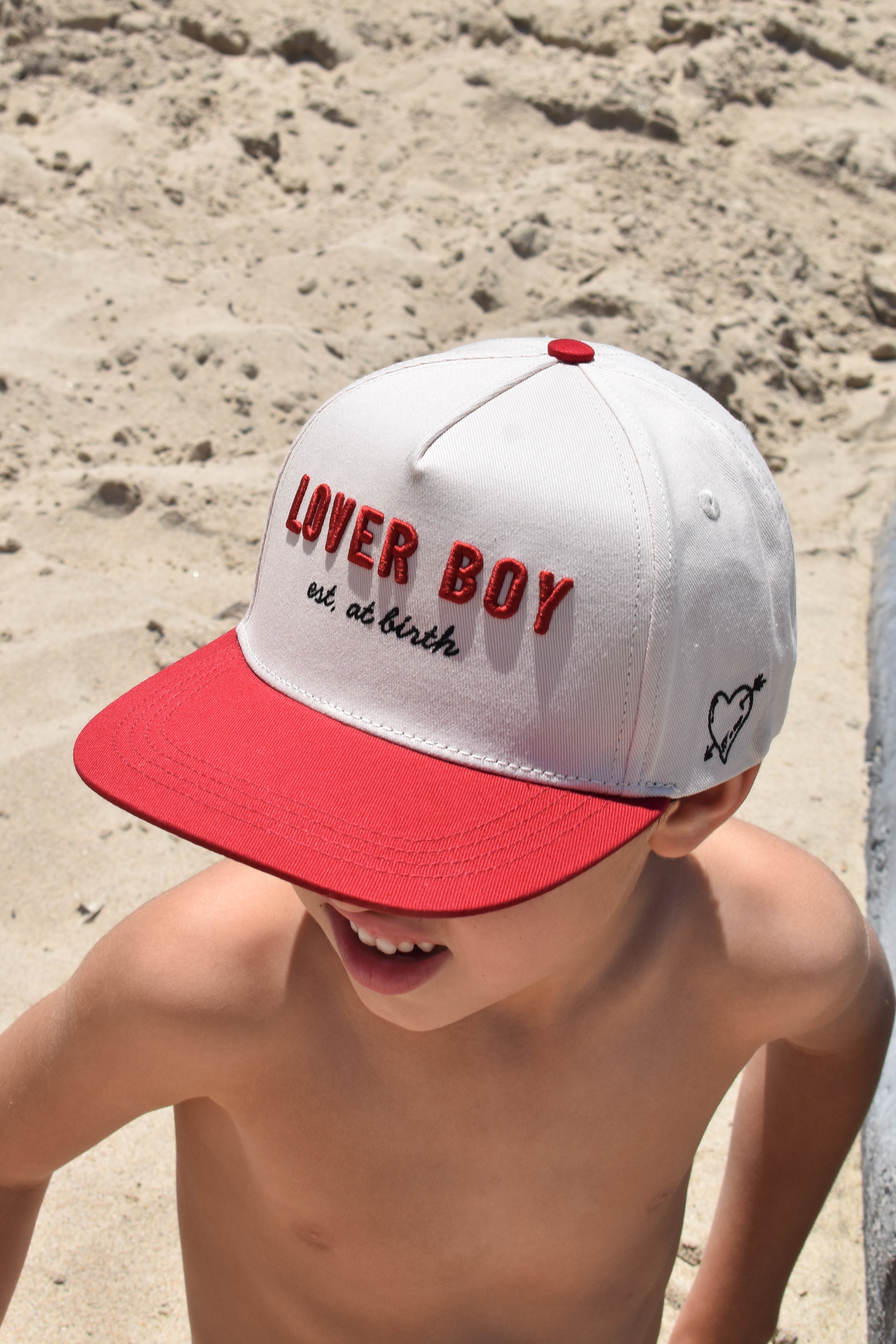 Child wearing a white and red cap with 'LOVER BOY' text on sandy ground