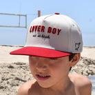 Child wearing a 'Liver Boy' cap on a beach with clear blue sky