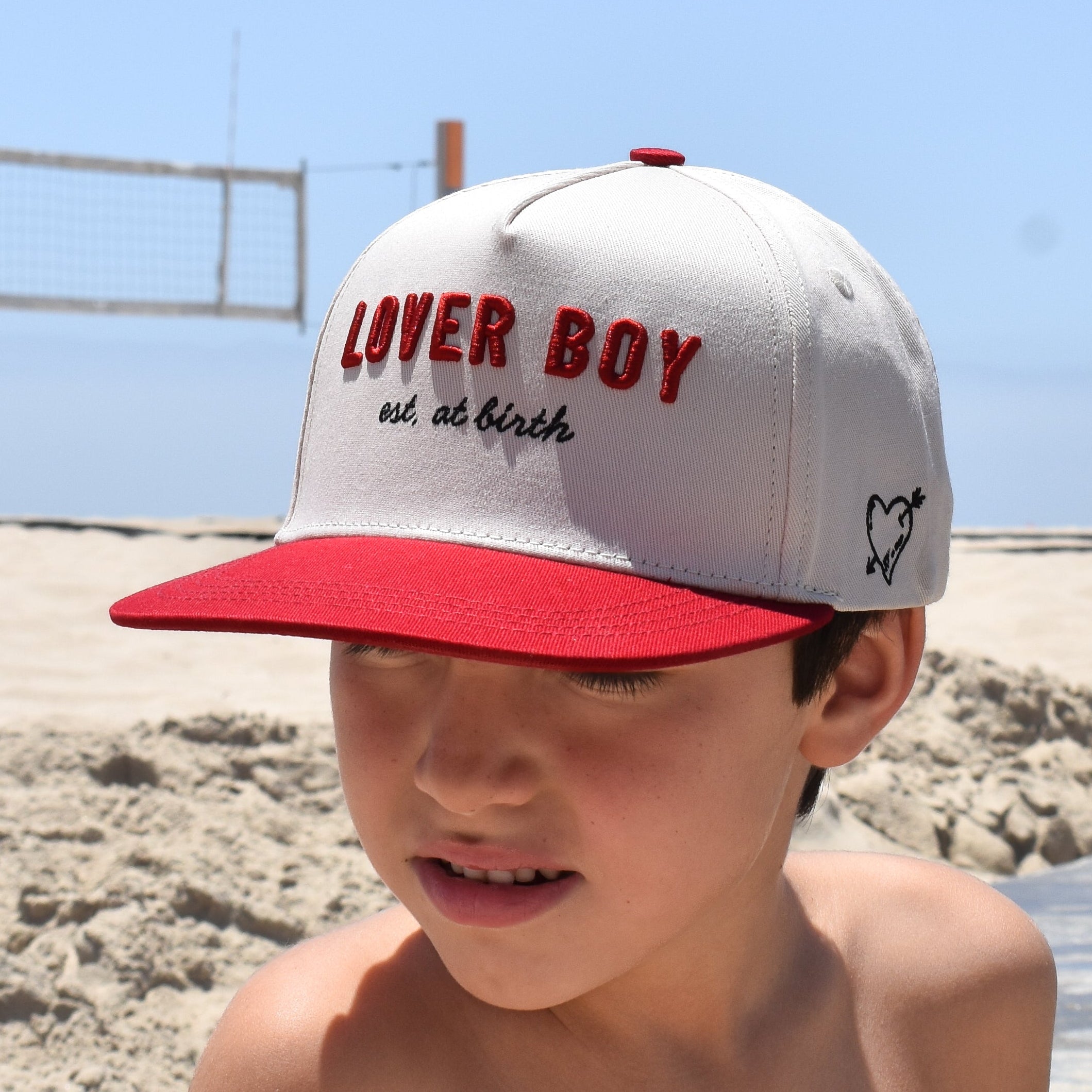 Child wearing a 'Liver Boy' cap on a beach with clear blue sky