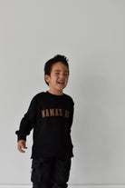 A young boy laughing while wearing a black crewneck sweatshirt with 'MAMAS BOY' printed in gold across the chest, paired with black cargo pants, set against a plain light gray wall.