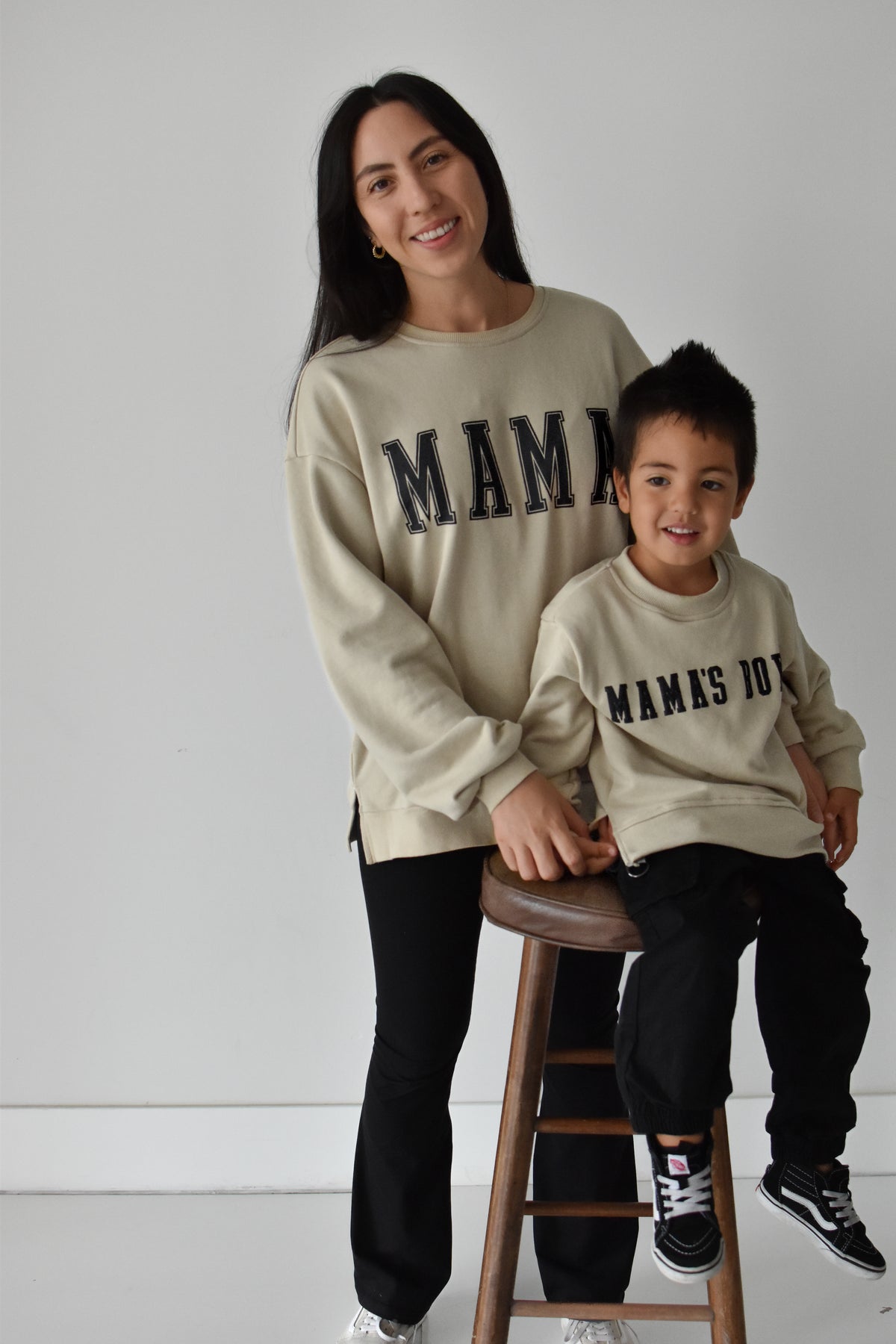 A woman and a young boy wearing matching beige crewneck sweatshirts with 'MAMA' and 'MAMAS BOY' printed in black varsity-style lettering, posed in a studio setting with a wooden stool.