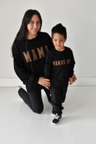 A woman and a young boy wearing matching black sweatshirts with gold 'MAMA' and 'MAMA'S BOY' text, posing together in a bright white studio setting.