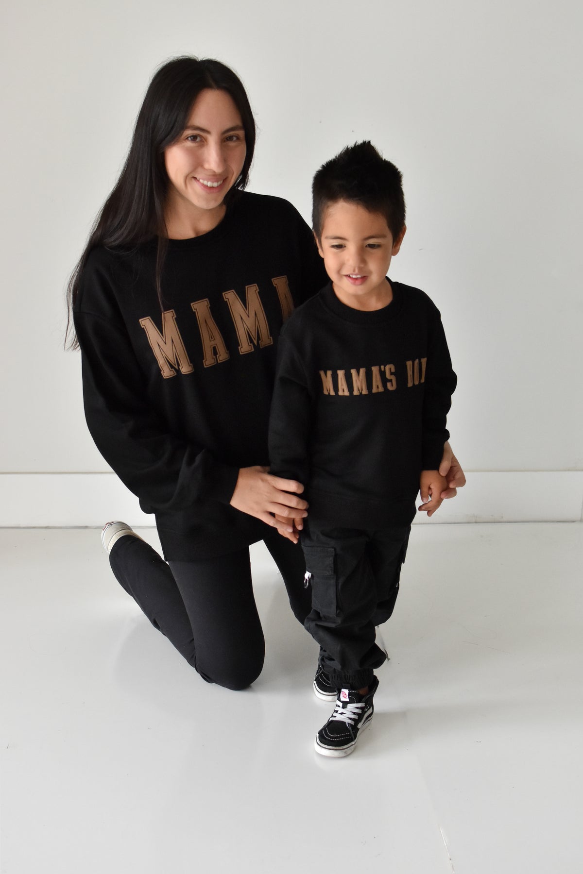 A woman and a young boy wearing matching black sweatshirts with gold 'MAMA' and 'MAMA'S BOY' text, posing together in a bright white studio setting.