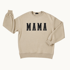 A beige crewneck sweatshirt featuring the word 'MAMA' printed across the chest in black varsity-style block letters with white outlines, displayed on a clean off-white background.