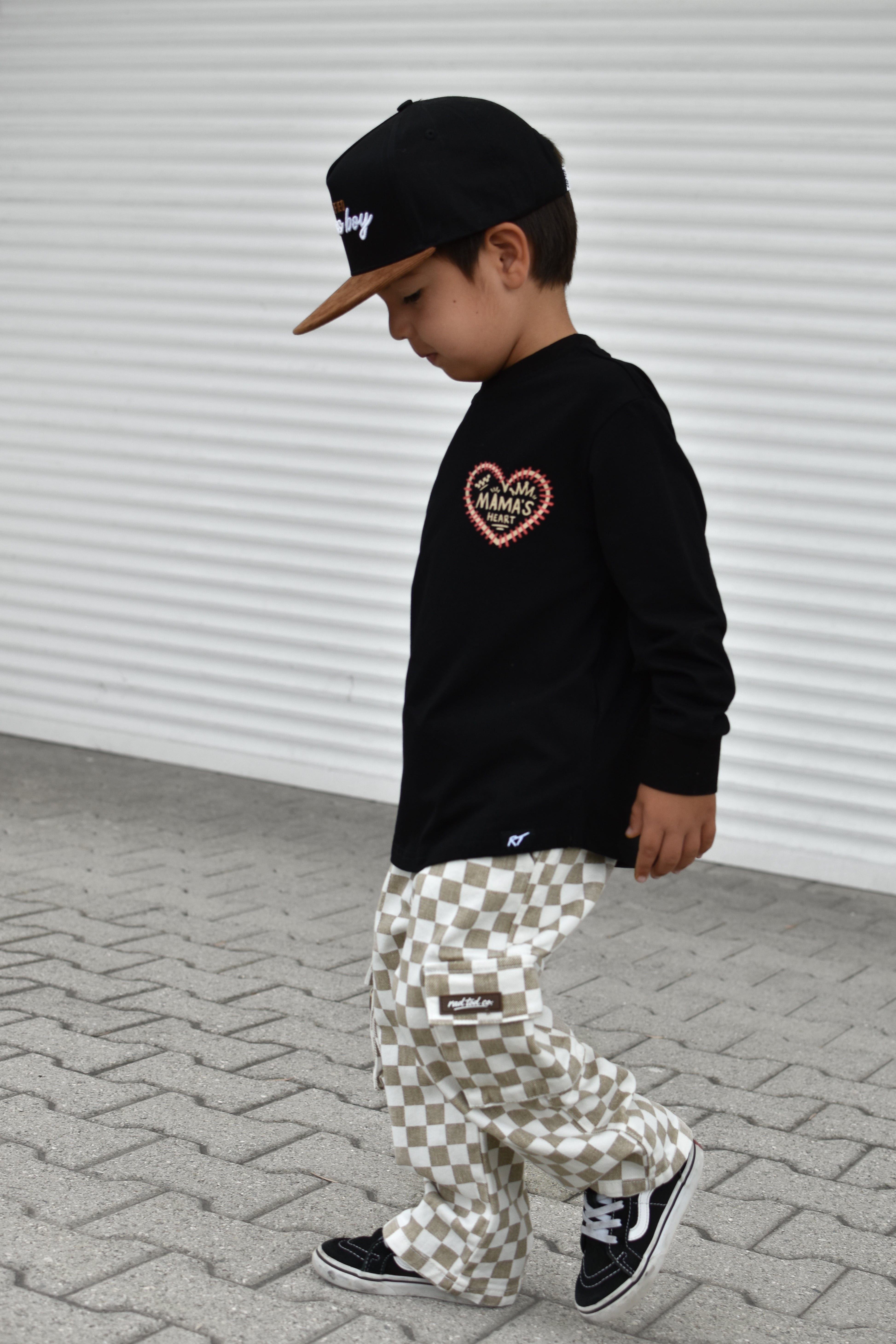 A child wearing a black long-sleeve shirt with a heart graphic, beige and white checkered pants, and a black cap, walking on a paved surface against a white corrugated wall.