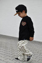 A child wearing a black long-sleeve shirt with a heart graphic, beige and white checkered pants, and a black cap, walking on a paved surface against a white corrugated wall.