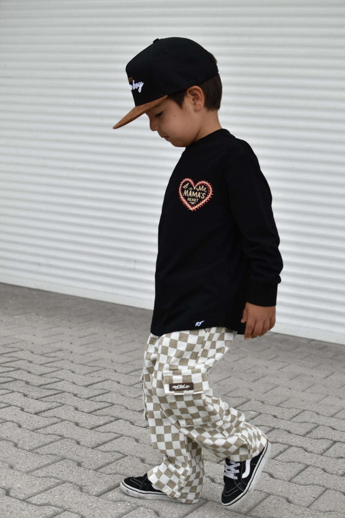 A child wearing a black long-sleeve shirt with a heart graphic, beige and white checkered pants, and a black cap, walking on a paved surface against a white corrugated wall.