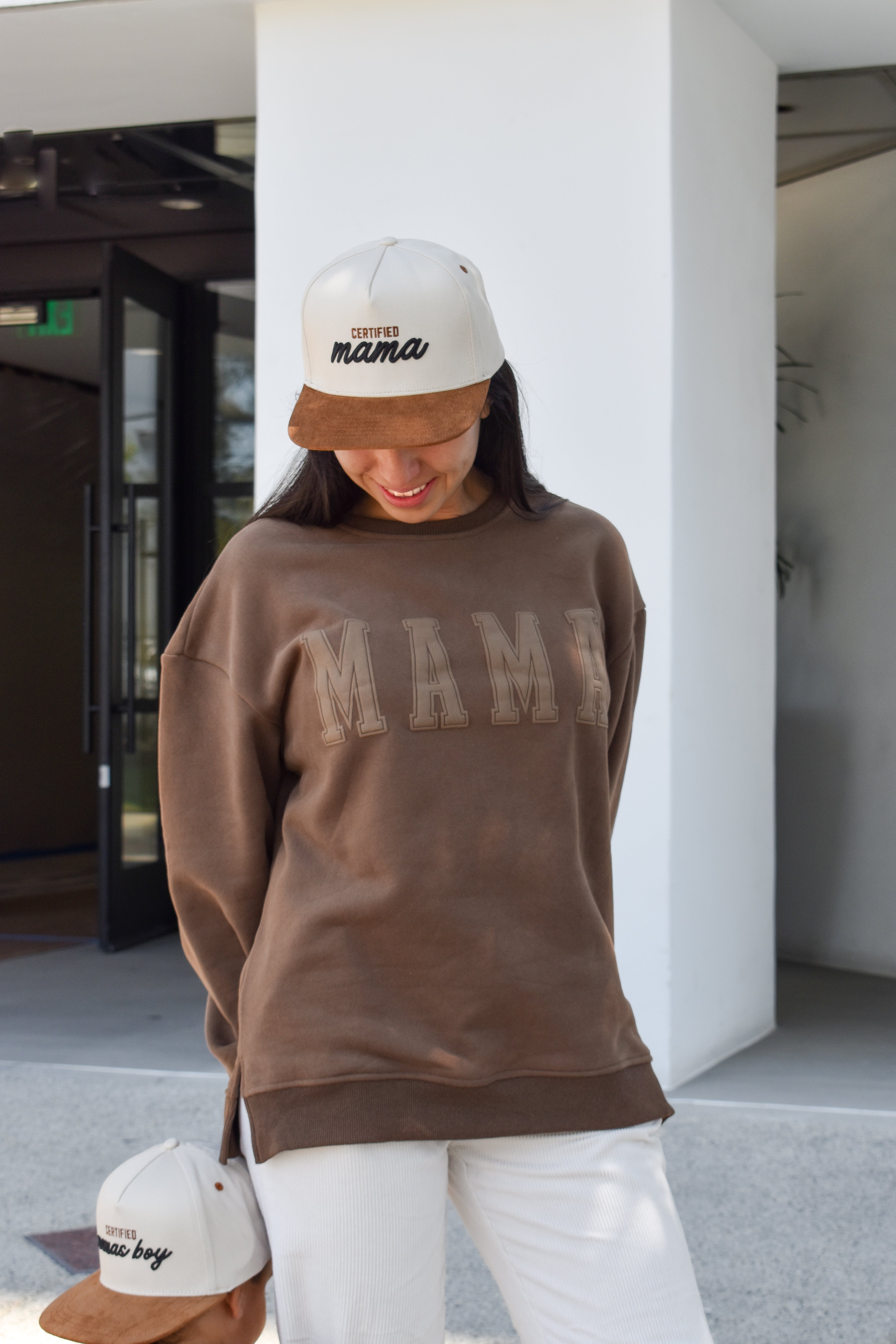 Person wearing a brown sweatshirt with 'MAMA' text, white pants, and a white cap 