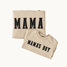 Two folded beige crewneck sweatshirts on a light off-white background. The larger sweatshirt in the back has 'MAMA' printed across the chest in black varsity-style block letters, and the smaller sweatshirt in the front has 'MAMA'S BOY' printed in a similar black font.
