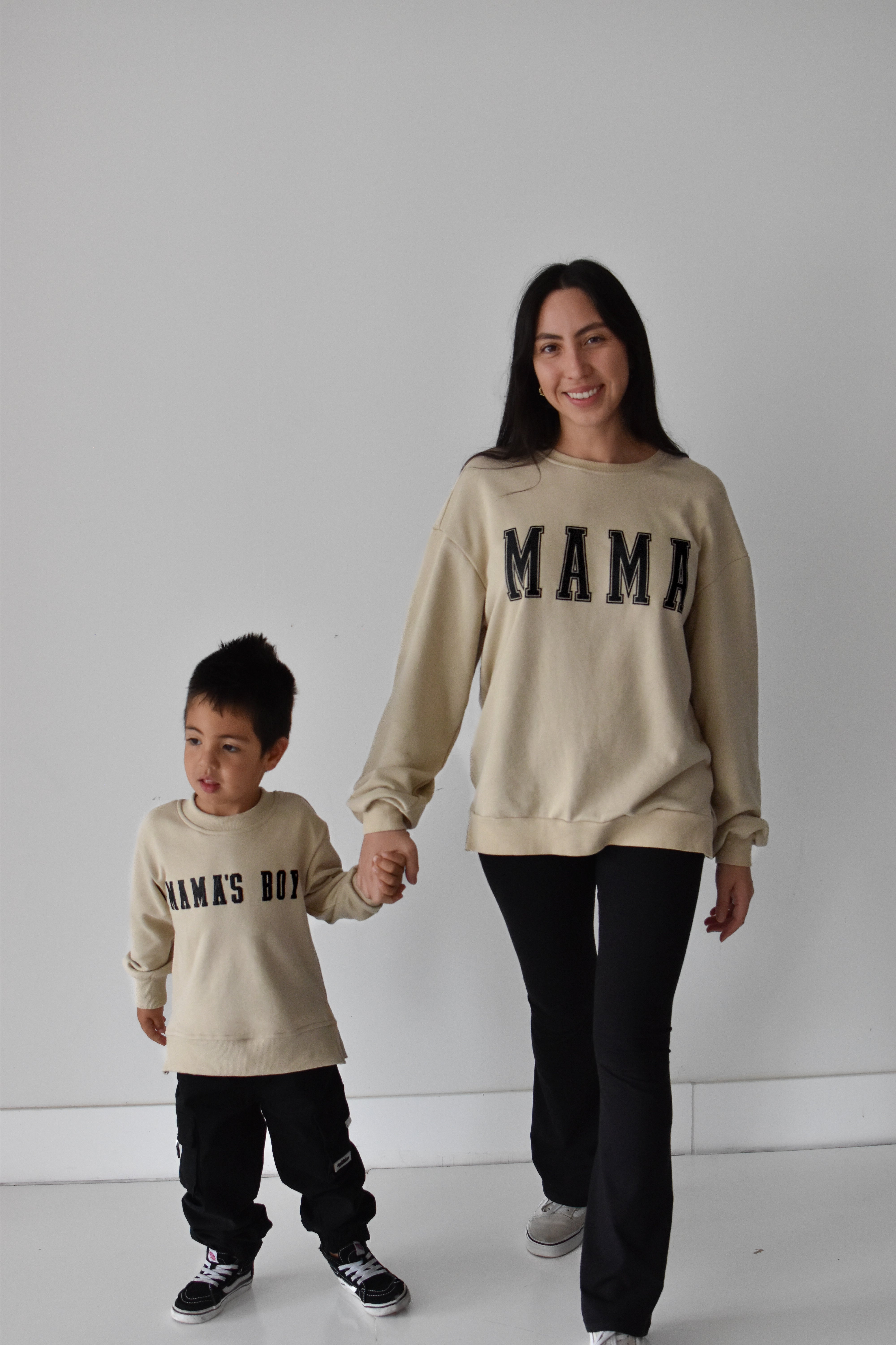 A woman and a young boy holding hands, both wearing matching beige crewneck sweatshirts with black lettering. The woman's sweatshirt reads 'MAMA' and the boy's reads 'MAMA'S BOY'.