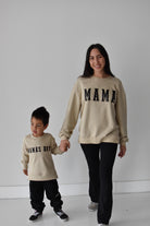 A woman and a young boy holding hands, both wearing matching beige crewneck sweatshirts with black lettering. The woman's sweatshirt reads 'MAMA' and the boy's reads 'MAMA'S BOY'.