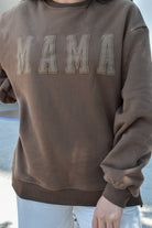 Brown sweatshirt with 'MAMA' text worn by a person outdoors.
