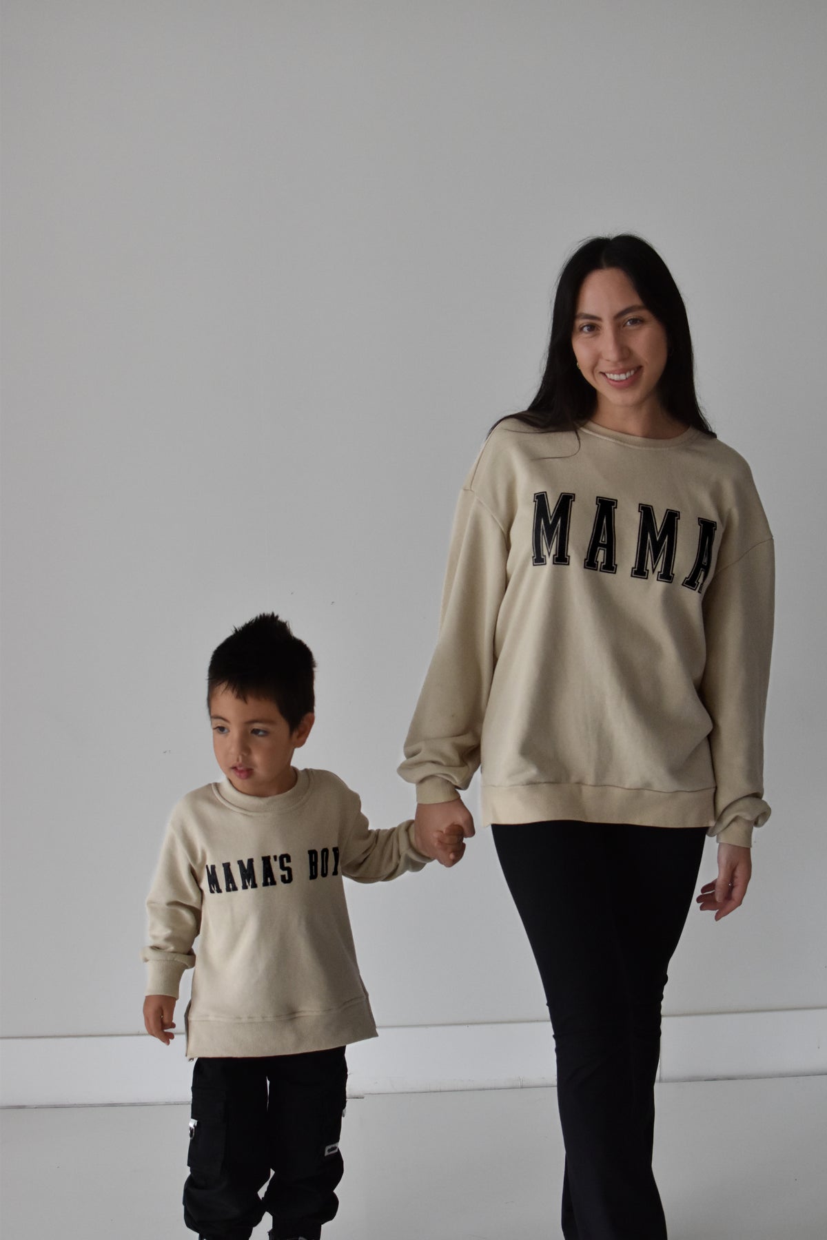 A woman and a young boy holding hands, wearing matching beige crewneck sweatshirts with black text. The woman's sweatshirt reads 'MAMA' and the boy's reads 'MAMAS BOY'.