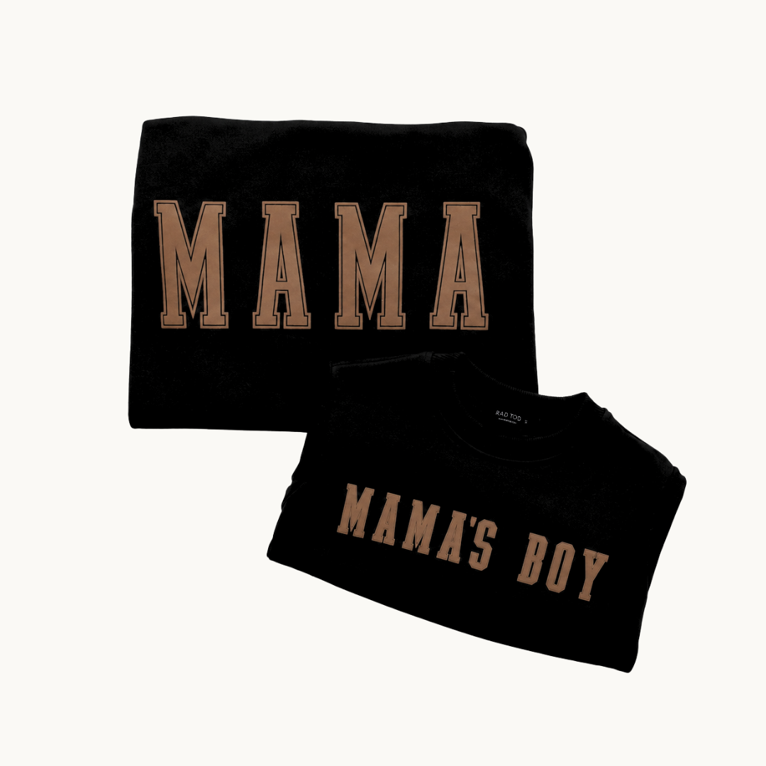 Two folded black crewnecks on a light cream background. The larger crewneck says 'MAMA' in tan varsity-style letters, and the smaller shirt says 'MAMA'S BOY' in the same tan lettering.