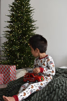 A young child wearing a white Christmas-themed pajama set with a holiday print sits on a dark green textured blanket in front of a lit Christmas tree, holding a red toy car.