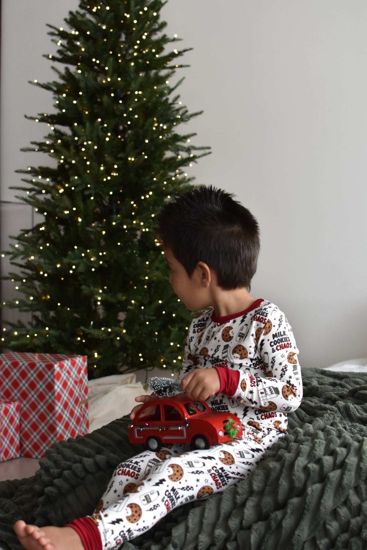 A young child wearing a white Christmas-themed pajama set with a holiday print sits on a dark green textured blanket in front of a lit Christmas tree, holding a red toy car.