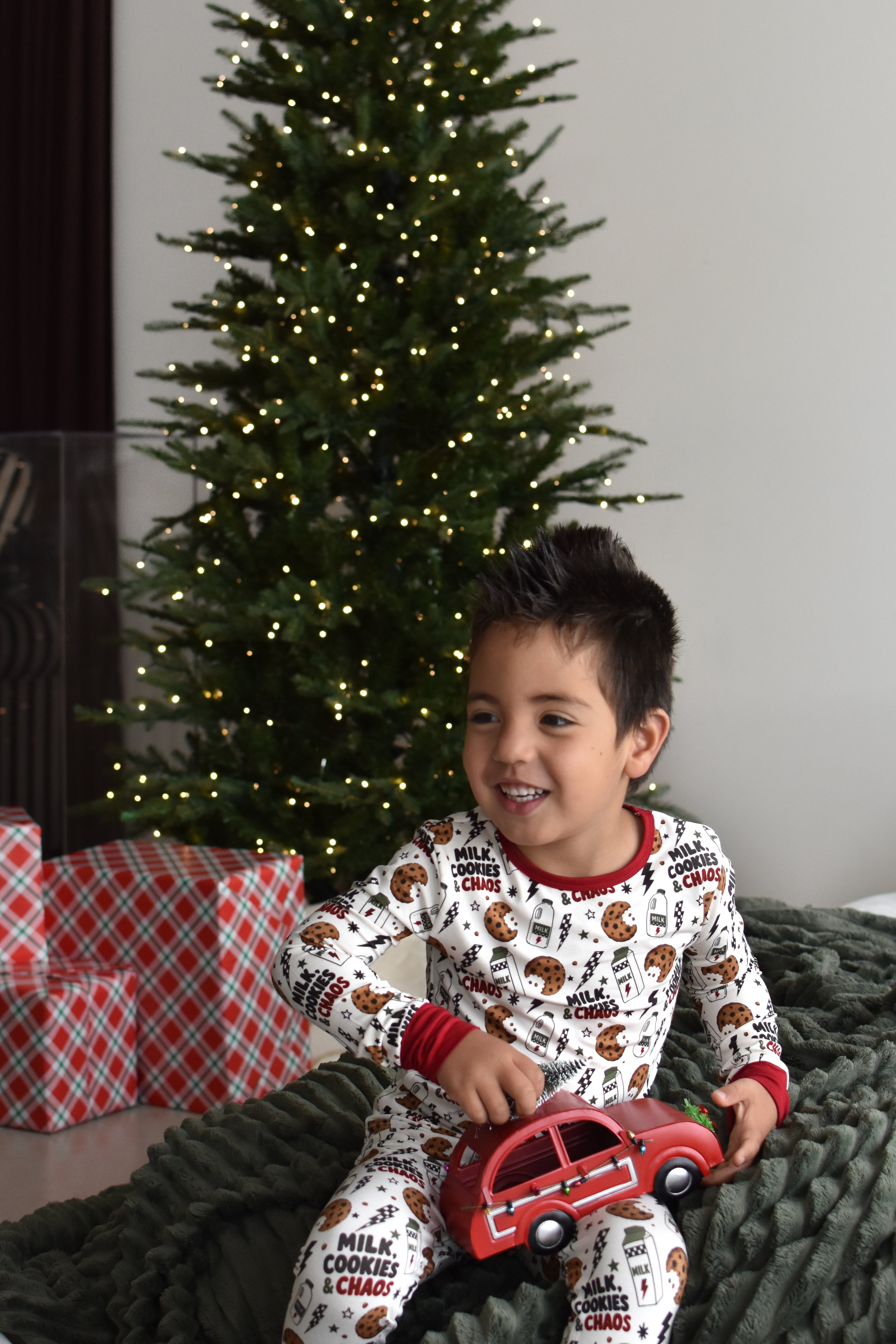 A young boy wearing a white and red 'Milk Cookies & Chaos' holiday pajama set sits on a dark green knit blanket in front of a decorated Christmas tree, holding a red toy car.