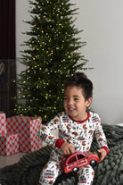 A young boy wearing a white and red 'Milk Cookies & Chaos' holiday pajama set sits on a dark green knit blanket in front of a decorated Christmas tree, holding a red toy car.