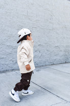 Child wearing a beige jacket, brown pants, and white sneakers against a gray wall.
