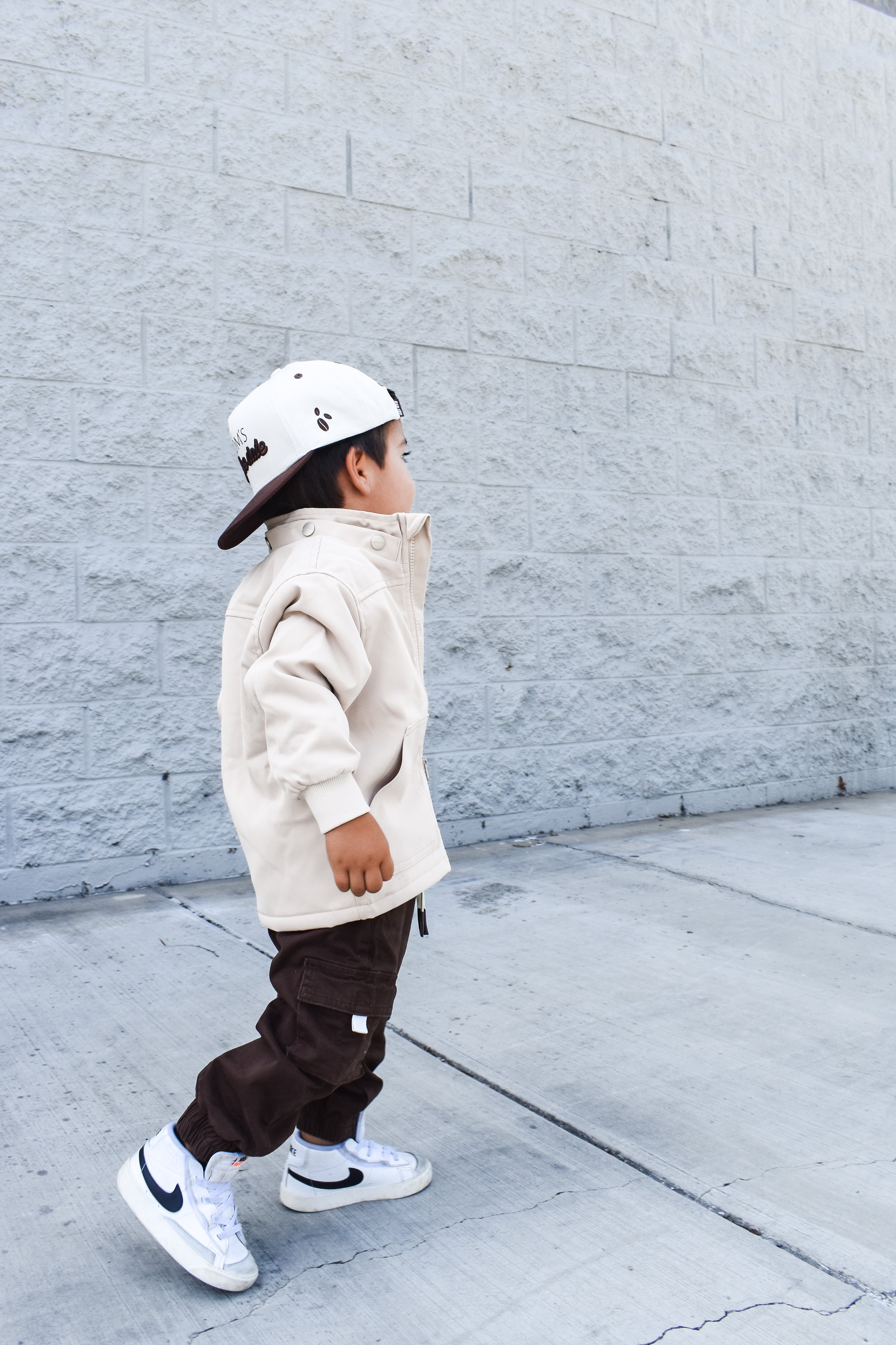 Child wearing a beige jacket, brown pants, and white sneakers against a gray wall.