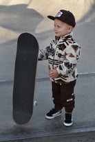 A young boy stands at a skatepark holding a skateboard, wearing a cream and brown patterned fleece jacket, dark brown pants, a black cap, and black sneakers.