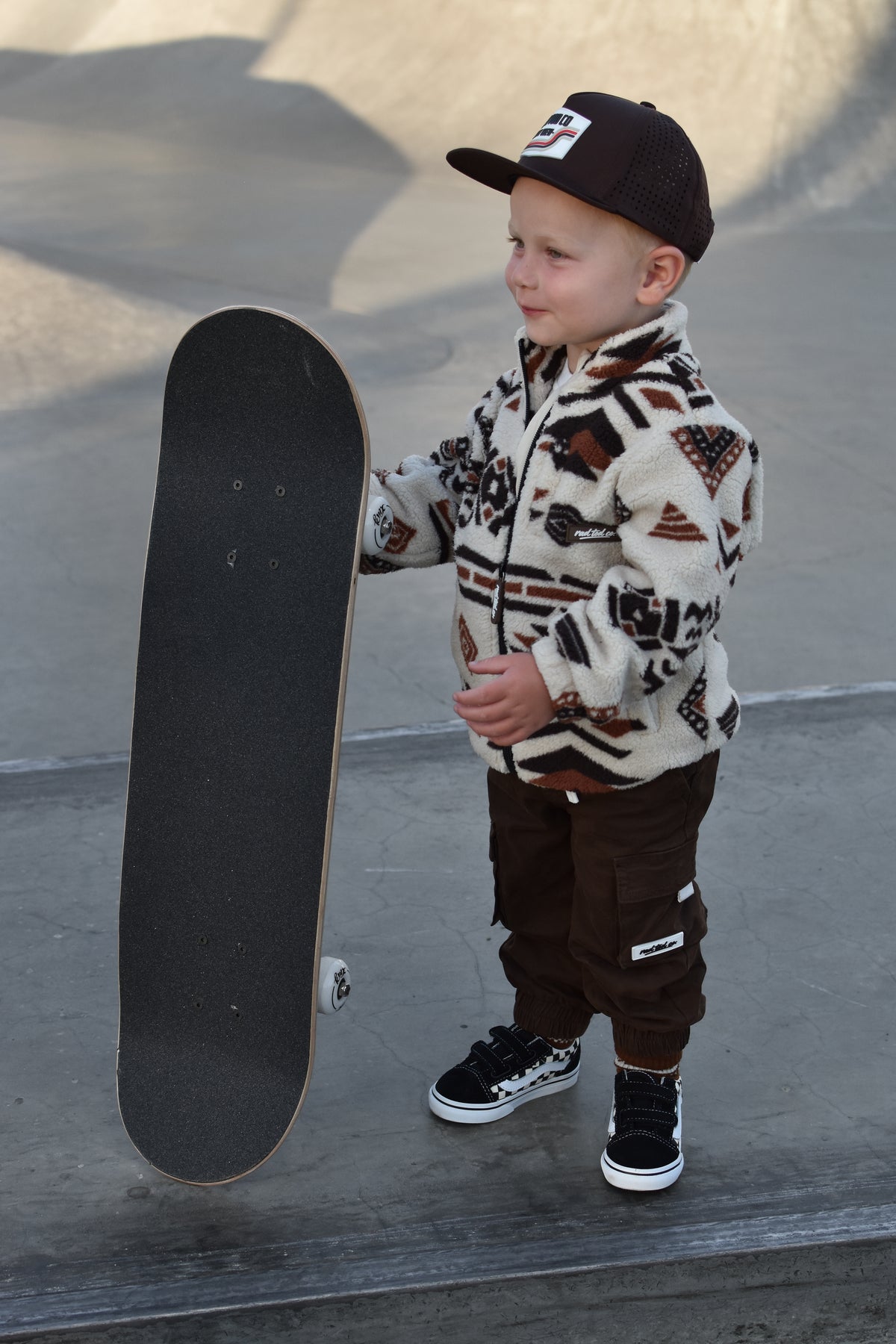 A young boy stands at a skatepark holding a skateboard, wearing a cream and brown patterned fleece jacket, dark brown pants, a black cap, and black sneakers.