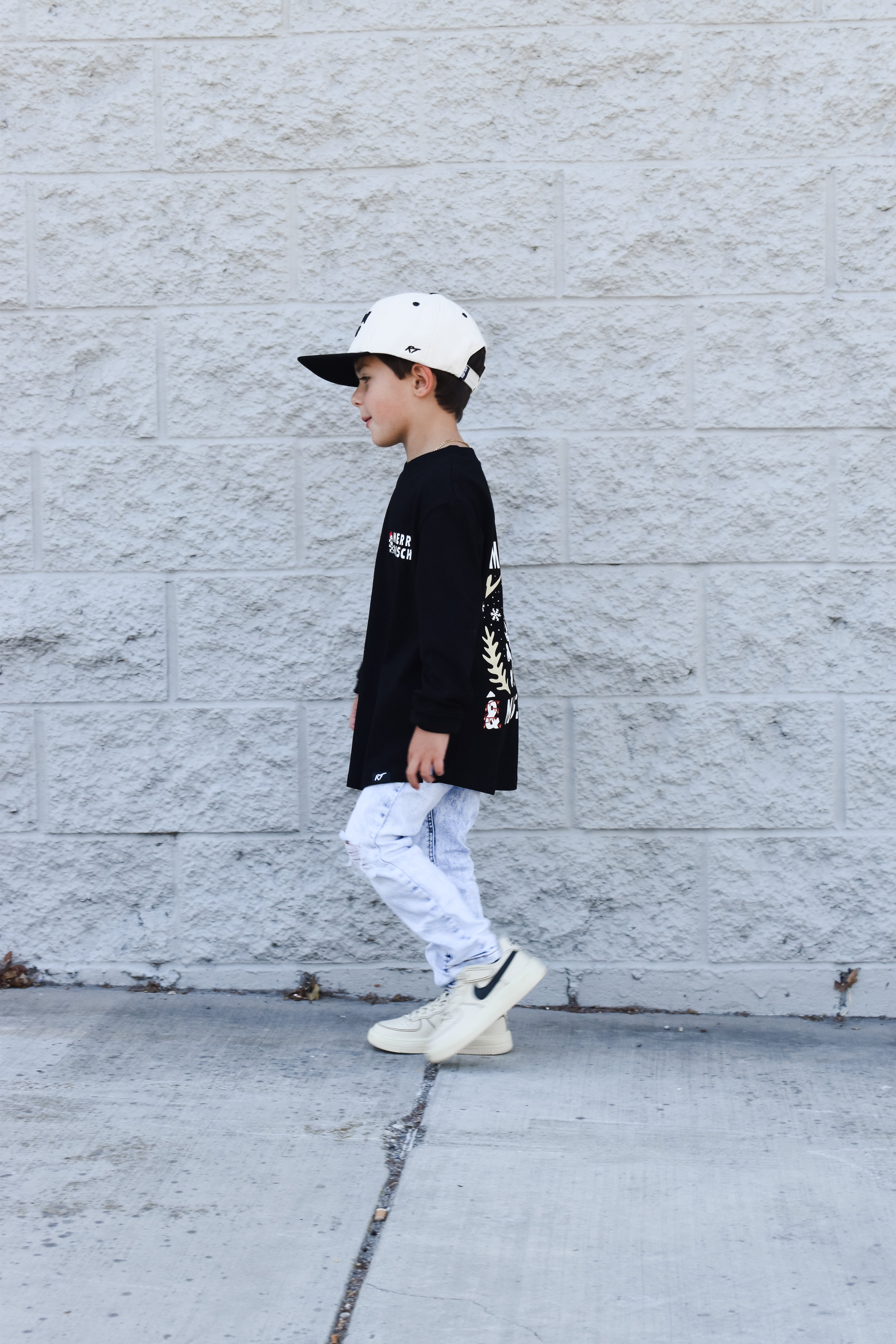 Child wearing a black long-sleeve shirt with white text, light blue jeans, and white sneakers against a gray wall.