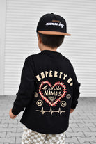A child shown from behind wearing a black long-sleeve shirt with a large back graphic that reads 'PROPERTY OF MAMA'S HEART' in cream and red, along with a black cap that says ' Certified mamas boy'.