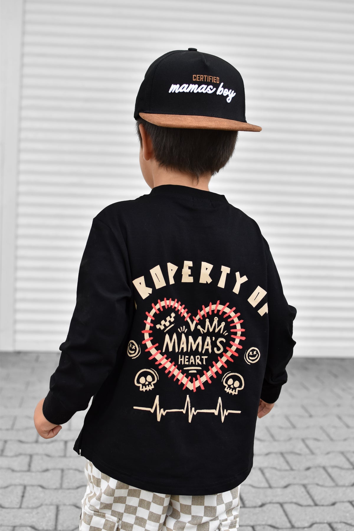 A child shown from behind wearing a black long-sleeve shirt with a large back graphic that reads 'PROPERTY OF MAMA'S HEART' in cream and red, along with a black cap that says ' Certified mamas boy'.