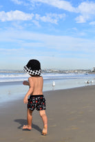 Child walking on a beach with a checkered hat and patterned shorts, facing the ocean.