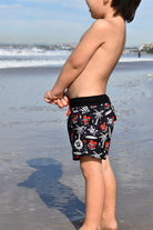 Toddler wearing Mama’s Little Surfer Swimmie in black with red heart and surf graphics at the beach